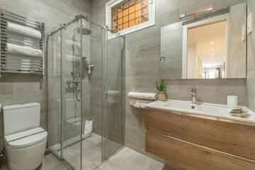 Bathroom with faux marble tile, walk-in shower with sliding glass doors, frameless mirror on the wall and vanity with white porcelain sink and white dresser and flower vase