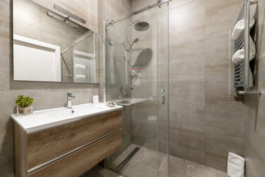 Bathroom with imitation marble tiles, shower cabin with sliding glass doors, frameless mirror on the wall and chest of drawers with white porcelain sink