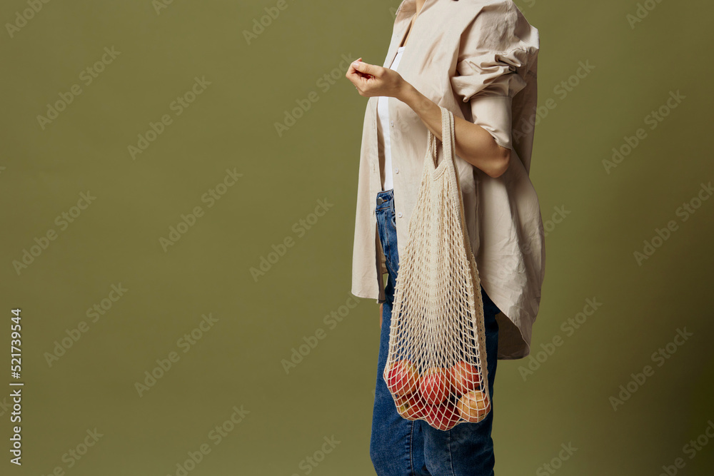 Wall mural Cropped shot of beautiful slim female in linen casual shirt hold string bag with fruit on elbow posing isolated on over olive green pastel background. Eco-friendly Natural products concept. Side view