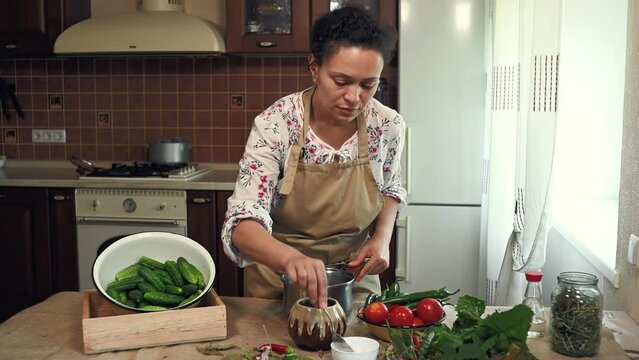 Multiethnic pleasant woman, housewife wearing chef apron, pouring salt and sugar into pot, adding ingredients while preparing pickle marinade for marinating seasonal vegetables in the home kitchen