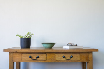 Small succulent plant in pot with green ceramic bowl, book and glasses on oak side table against beige wall (selective focus)