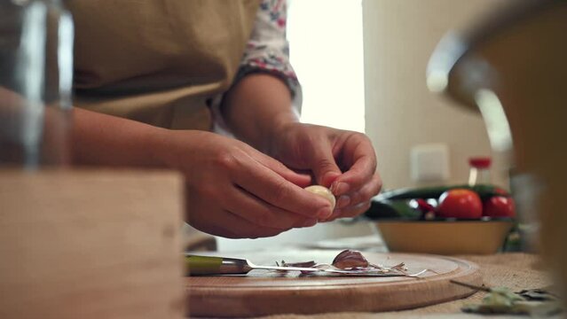 Details: Hands of a housewife in chef apron, peeling garlic by hands, at home kitchen. Woman putting dry bay leaf and peppercorns on a wooden chopping board, ingredients for preparing pickle