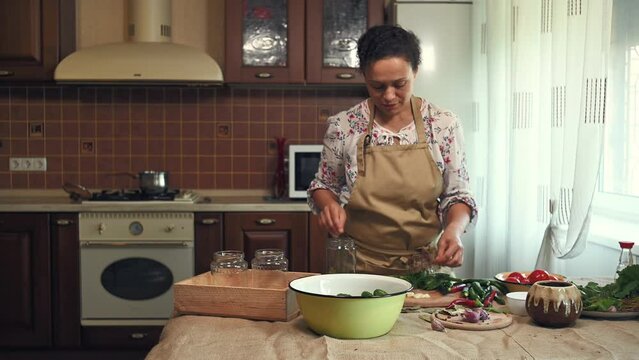 African American pretty woman, housewife in chef's apron, standing at the kitchen table and filling a sterilized glass jar with chopped dill leaves, garlic while marinating vegetables for the winter