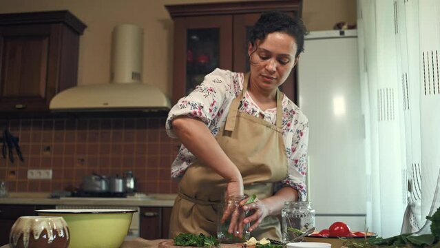 Pleasant multiethnic woman in chef apron, standing by kitchen table with ingredients for pickling and putting chopped fresh dill leaves and garlic cloves into a sterilized glass jar. Homemade canning