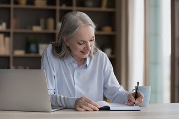 Happy focused mature freelancer woman working at laptop from home, writing notes in notebook,...