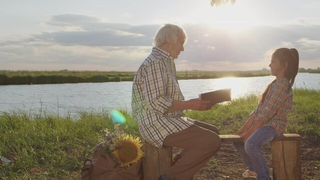 Happy smiling family spend time together outdoor : old caring grandmother read book story granddaughter at sunset. Relationship harmony of grandparents and children, imagination fantasy, peaceful life
