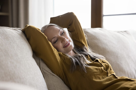 Happy relaxed beautiful mature woman resting on home sofa, leaning on back, looking away, enjoying relaxation, leisure, comfort, dreaming, smiling at good thoughts, breathing fresh air, watching TV