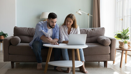 Serious millennial couple sit on sofa sorting out papers, reviewing bills for utility expenses,...
