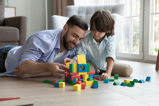 Father lying on warm floor in living room play with preschooler son use wooden multicoloured blocks, construct castle look focused spend pastime together at modern home. Developmental games with kids