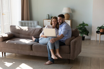 Obraz premium Couple sit on sofa use laptop make on-line purchase, order services, buy goods, search home decorations ideas on internet spend time with modern technology. E-commerce clients for comfort life concept
