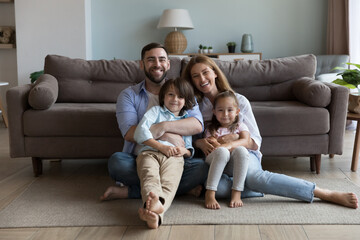 Couple with little children hugging sit on floor in living room smile look at camera feel happy, enjoy new house and weekend together at own first home. Bank loan, well-being family portrait concept