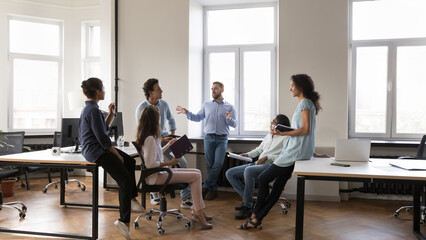 Multiethnic staff members gathered in coworking solve business, listen to male team leader during morning briefing in modern company office. Boss share information, corporate goals, tell about project