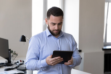 Man, office employee stand at workplace working on digital tablet, contact to client by e-mail app use wireless device, prepare on-line presentation. Workflow, lifestyle, modern tech, business concept