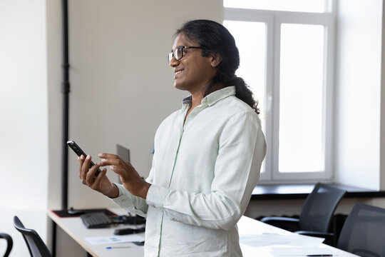 Thoughtful Indian man entrepreneur standing in office with smartphone smile, enjoy easy comfort electronic services during break at modern workplace, use business mobile application, lifestyle concept