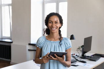 Obraz premium Young 35s African businesslady pose at workplace with digital tablet in hands smile look at camera. Workflow use modern wireless tech, apps, remote communication to client, corporate messaging concept