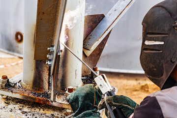 An elderly gray-haired gray-haired welder assembles metal structures at a construction site. Arc welding work. Sparks from welding. Close-up.