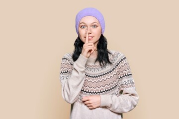Young female in sweater hat showing finger gesture secret, shh, on brown background