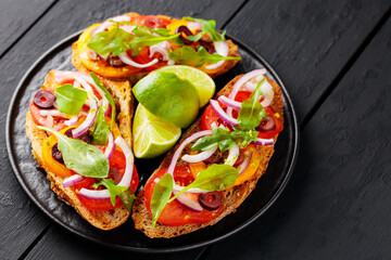 Healthy vegetarian toasts and lime on a black plate. Bruschetta with tomatoes, greens and olives. Healthy open sandwiches on black boards. Copy space. Top view