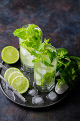 Two glasses of mojito cocktail, lime slices, mint and ice on dark background. Refreshing mint cocktail mojito, ice tongs and cocktail spoon on black plate