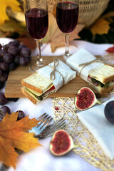 Close up of romantic picnic with sandwich, grapes and glasses red wine on background of autumn garden with maple leaves in evening outdoors. Still life