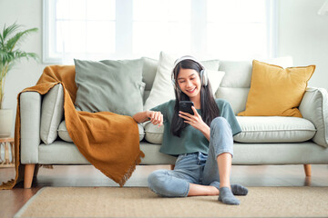 Happy asian woman listening to music from mobile phone while sitting on the rug beside to the sofa...