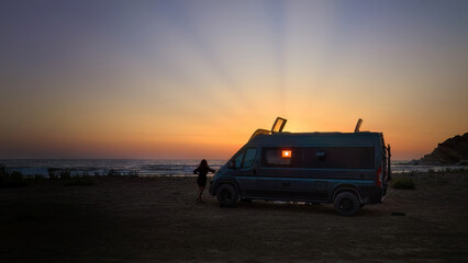 Fototapeta na wymiar Silhouette of a offroad caravan and a young woman against the setting sun, the beach and the sea. Adventure of wild camping, nomadic life. Travelling in a van camper.