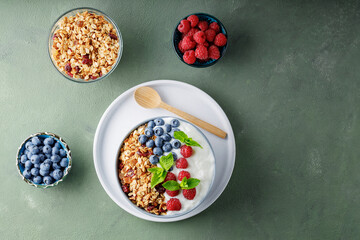 Obraz na płótnie Canvas Greek yogurt with raspberries, blueberries and granola on a green background. Bowl with healthy breakfast on white plate with wooden spoon. Top view. Copy space