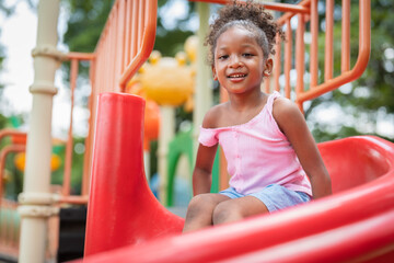 Smiling African American little girl with curly hair has fun sitting on slider at playground in the...