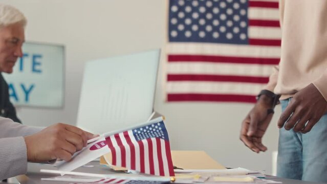 Midsection slowmo of electoral district worker giving US flag, sticker and bulletin to unrecognizable Black man came to vote on Elections Day