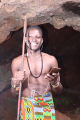 A cave in Africa: Masai Moran warrior with a spear smiling listening to music on a mobile phone...