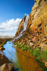 Waterfall in a granite quarry, landscape with red stones and blue sky. Granite mining.