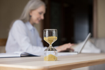 Obraz na płótnie Canvas Hourglass with flowing sand on work desk of elderly businesswoman, closed up object. Mature business professional lady typing on laptop in blurred background. Career, time management concept