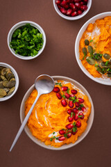 Pumpkin puree soup of butternut squash in bowls with pumpkin and pomegranate seeds and green onions, top view on plates with food on brown background