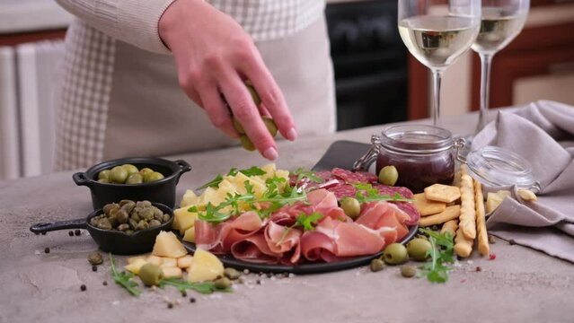 meat and cheese plater at domestic kitchen - woman puts olives to serving board with sausages, cheese and prosciutto ham