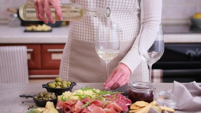 woman pouring wine to a glass from a bottle at domestic kitchen with meat and cheese plater on the table