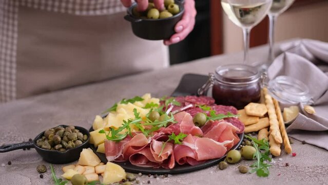 meat and cheese plater at domestic kitchen - woman puts olives to serving board with sausages, cheese and prosciutto ham