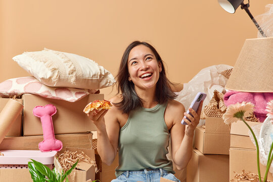 Positive young Asian woman with dark hair eats pizza holds smartphone focused above gladfully dressed in casual clothes going to relocate in new apartment or house surrounded by cardboard boxes.