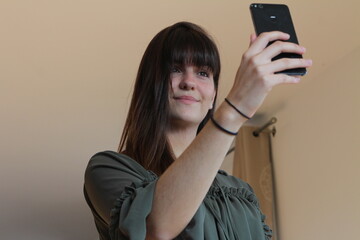 Girl taking a selfie with her new haircut. Young woman with bangs haircut smiling at the camera. Girl holding her phone. Green shirt.