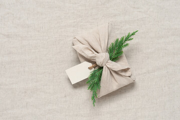 Christmas gift wrapped in fabric with juniper, beige linen textile background. A traditional Japanese furoshiki gift. Zero waste concept. Top view, selective focus, flat lay