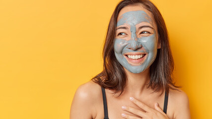 Horizontal shot of positive Asian woman with dark hair applies facial mud mask to reduce pores smiles happily looks away keeps hand on chest isolated over yellow background empty space for promo