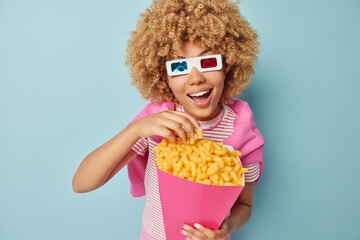 Obraz premium Surprised cheerful woman with curly bushy hair watches movie wears cinema eyeglasses eats corn sticks dressed in casual clothes isolated over blue background. Female model enjoys favorite snack
