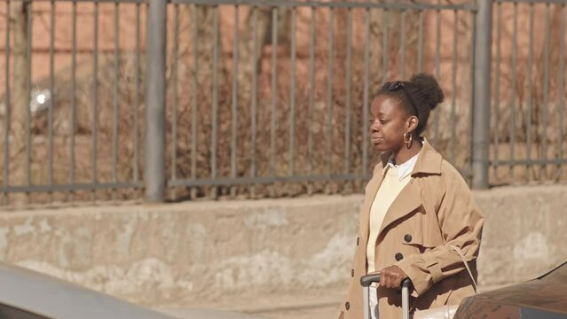 Medium slowmo of young Black woman in camel trench coat with suitcase catching cab outdoors on sunny day