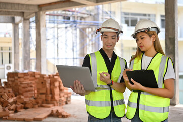 Portrait of Two Construction Engineers or Architects wearing white safety helmet and reflective clothing discussing at work with laptop on construction site.