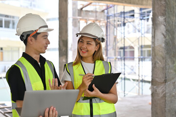 Portrait of Two Construction Engineers or Architects wearing white safety helmet and reflective clothing discussing at work with laptop on construction site.