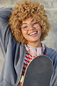 Youth lifestyle and recreation concept. Glad pretty teenage girl smiles toothily poses with skateboard rests after skating wears grey sweatshirt and spectacles uses headphones for listening music