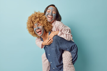 Horizontal shot of cheerful women give piggyback have fun foolish around apply beauty facial mask before going to bed dressed in slumber suit isolated over blue background. Friends spend time together