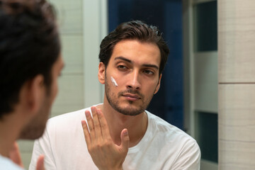 Handsome man looking at mirror and applying moisturizing cream on cheeks in bathroom. Groomed young guy doing skincare morning routine after taking a shower