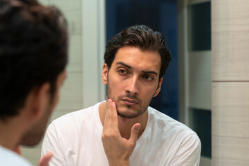 Fototapeta na wymiar Handsome man looking at mirror and applying moisturizing cream on cheeks in bathroom. Groomed young guy doing skincare morning routine after taking a shower