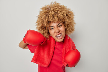 Strong curly female boxer wears mouthguard and boxing gloves red towel around neck practices fighting skills in gym being strict coach isolated over grey background ready to give nice punch.