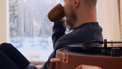 A bald man with a beard sits on the sofa, drinks coffee and listens to classical music, the man looks out the window with a beautiful view of the winter landscape. Record player for listening to music